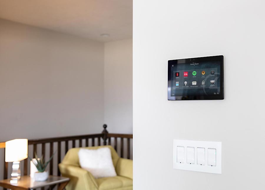 Control4 touchscreen mounted to the wall above a four-gang light switch. A sitting room is in the background.