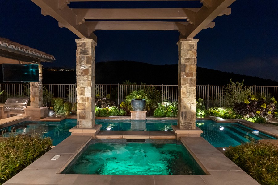Nighttime view of a luxurious outdoor pool and hot tub accented by landscape lighting and stone columns under a pergola.